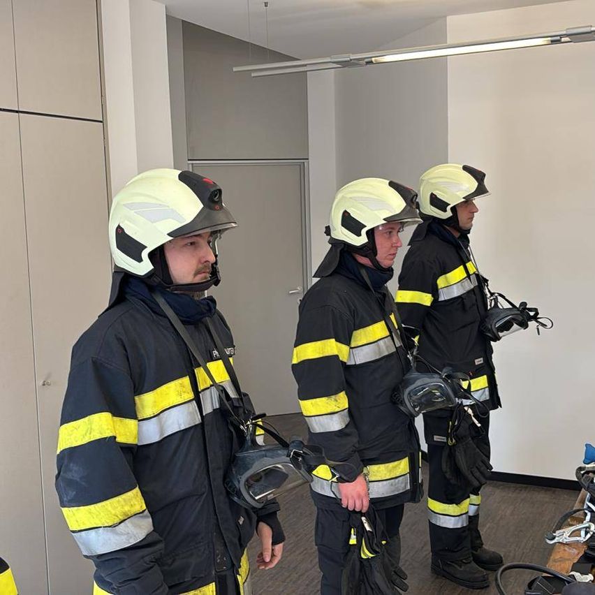 Drei Feuerwehrleute in Schutzkleidung stehen in einem Raum. Einer trägt einen Helm, ein anderer hat eine Maske. In der Nähe befinden sich Ausrüstungsgegenstände.