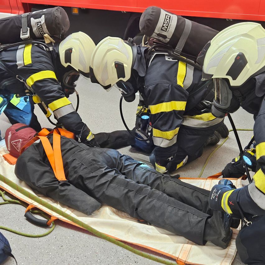 Eine Gruppe von Feuerwehrleuten in Schutzkleidung kümmert sich um eine Person, die auf einer Trage liegt. Ein Feuerwehrmann hält eine Maske, und die Person trägt einen Gurt.
