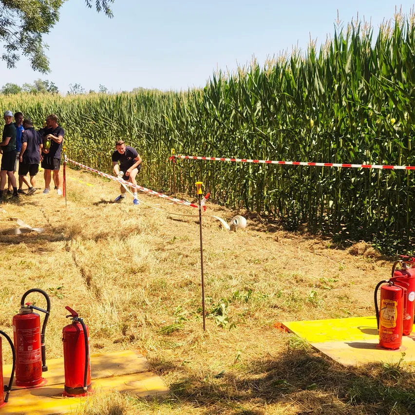 Eine Gruppe von Menschen steht in einem Maisfeld, ein Mann kickt einen Fußball. Es gibt Feuerlöscher und eine gelbe Matte auf dem Boden.