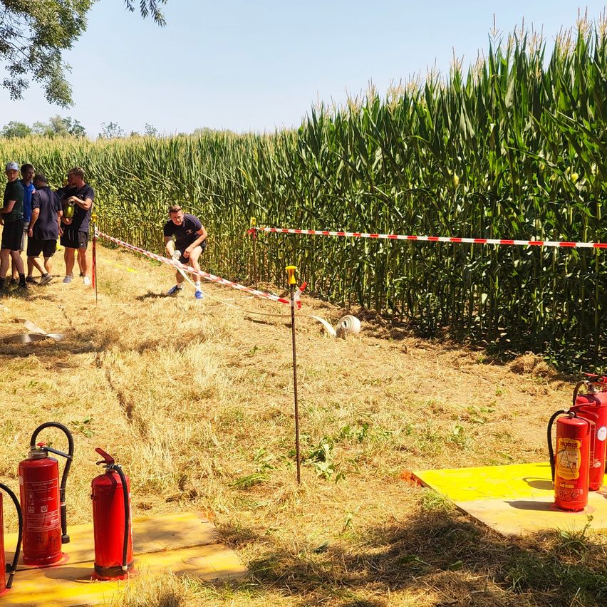 Eine Gruppe von Menschen steht in einem Maisfeld, ein Mann kickt einen Fußball. Es gibt Feuerlöscher und eine gelbe Matte auf dem Boden.