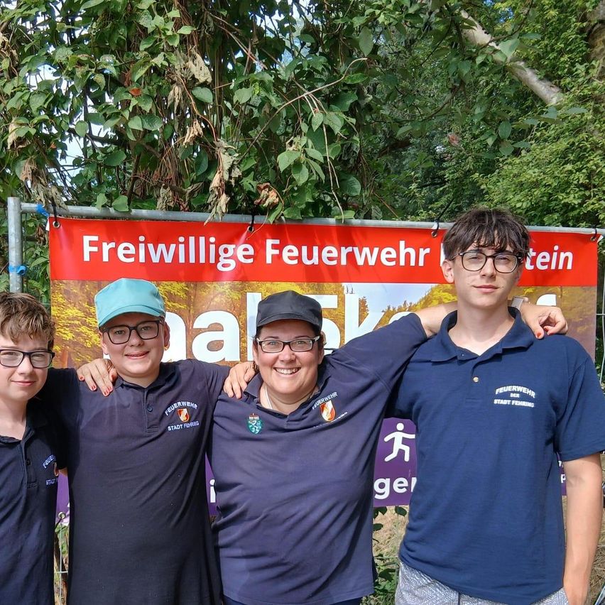 Drei junge Jungen und eine Frau stehen vor einem Banner mit der Aufschrift 'Freiwillige Feuerwehr'. Die Frau trägt eine Mütze und eine Brille, und die Jungen tragen Brillen und Poloshirts.