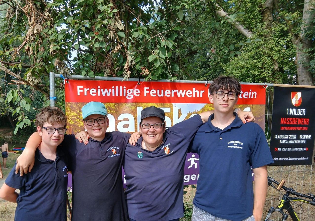 Drei junge Jungen und eine Frau stehen vor einem Banner mit der Aufschrift 'Freiwillige Feuerwehr'. Die Frau trägt eine Mütze und eine Brille, und die Jungen tragen Brillen und Poloshirts.