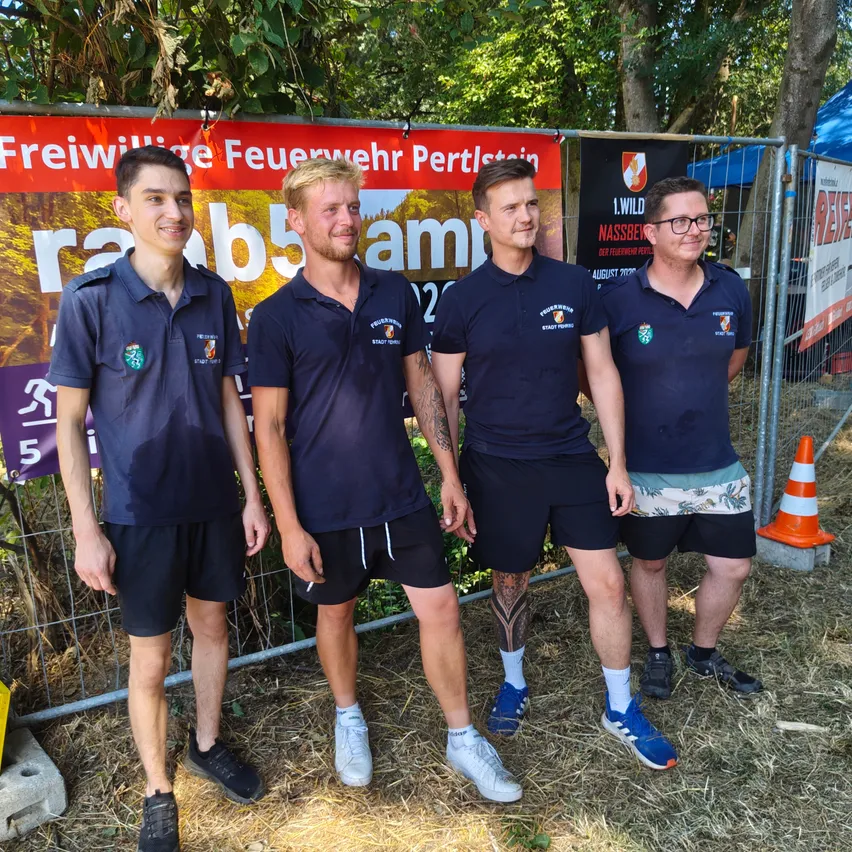 Vier Männer in Marineblau-Uniformen stehen zusammen draußen und lächeln für ein Foto. Sie stehen vor einem Zaun mit einem Banner und einem Verkehrskegel.