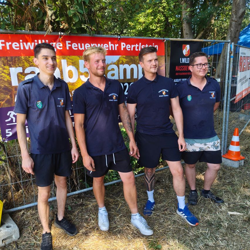 Vier Männer in Marineblau-Uniformen stehen zusammen draußen und lächeln für ein Foto. Sie stehen vor einem Zaun mit einem Banner und einem Verkehrskegel.