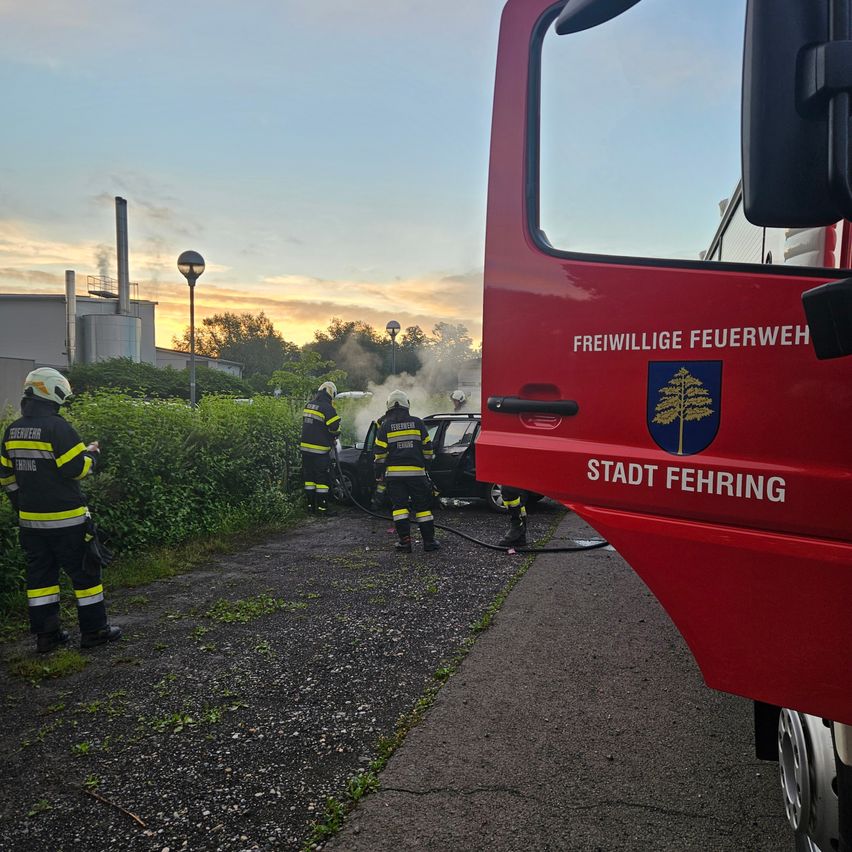 Eine Gruppe Feuerwehrleute reagiert auf einen Autobrand und benutzt eine Feuerwehrspritze, um die Flammen zu löschen. Das Fahrzeug ist am Straßenrand geparkt.