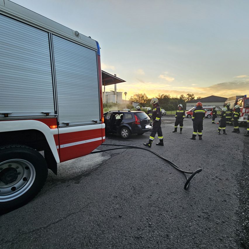 Feuerwehrleute in Uniform mit Helmen stehen um ein demoliertes Auto herum, in der Nähe eines Feuerwehrwagens und anderer Ausrüstung.