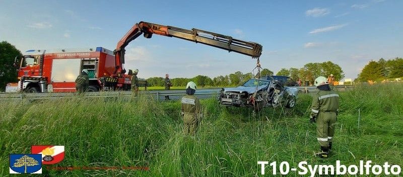 Ein Kran hebt ein demoliertes Auto von der Straße, Feuerwehrleute sind in der Nähe. Ein roter Feuerwehrwagen ist dahinter geparkt.