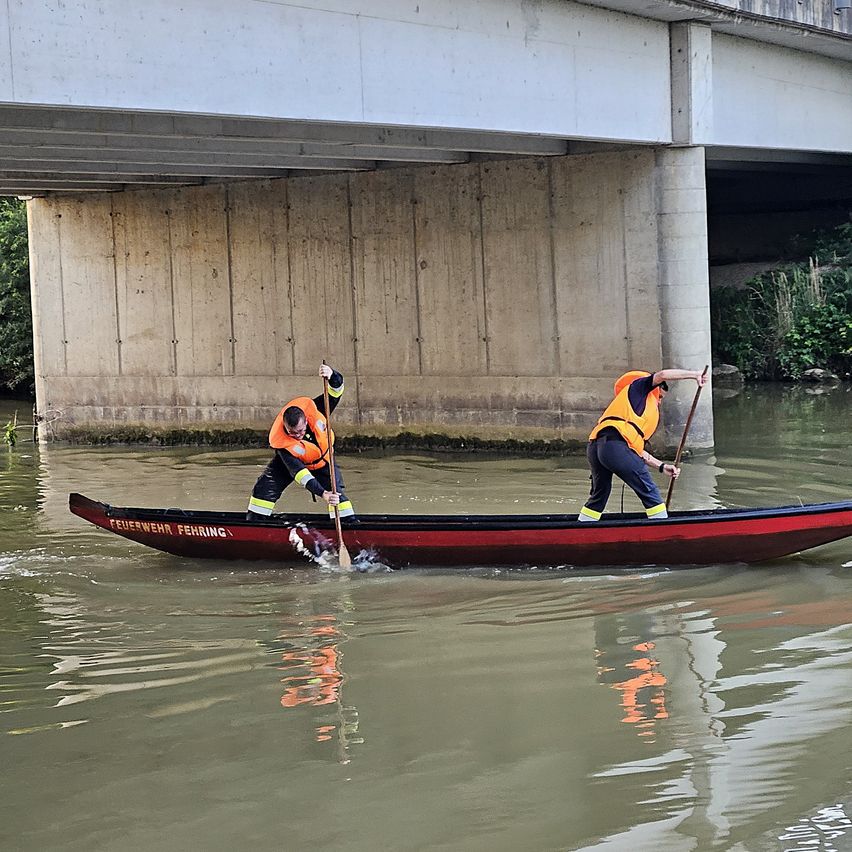 Zwei Personen in Sicherheitswesten rudern mit einem roten Boot unter einer Brücke. Das Wasser ist ruhig, und das Boot trägt den Text 'Feuerwehr Fährung'.