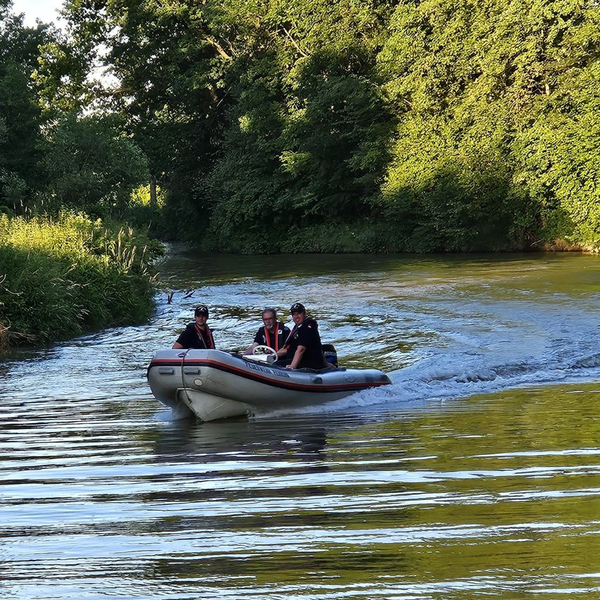 Drei Personen in einem Boot auf einem Fluss mit Bäumen auf beiden Seiten unter einem hellen Himmel.