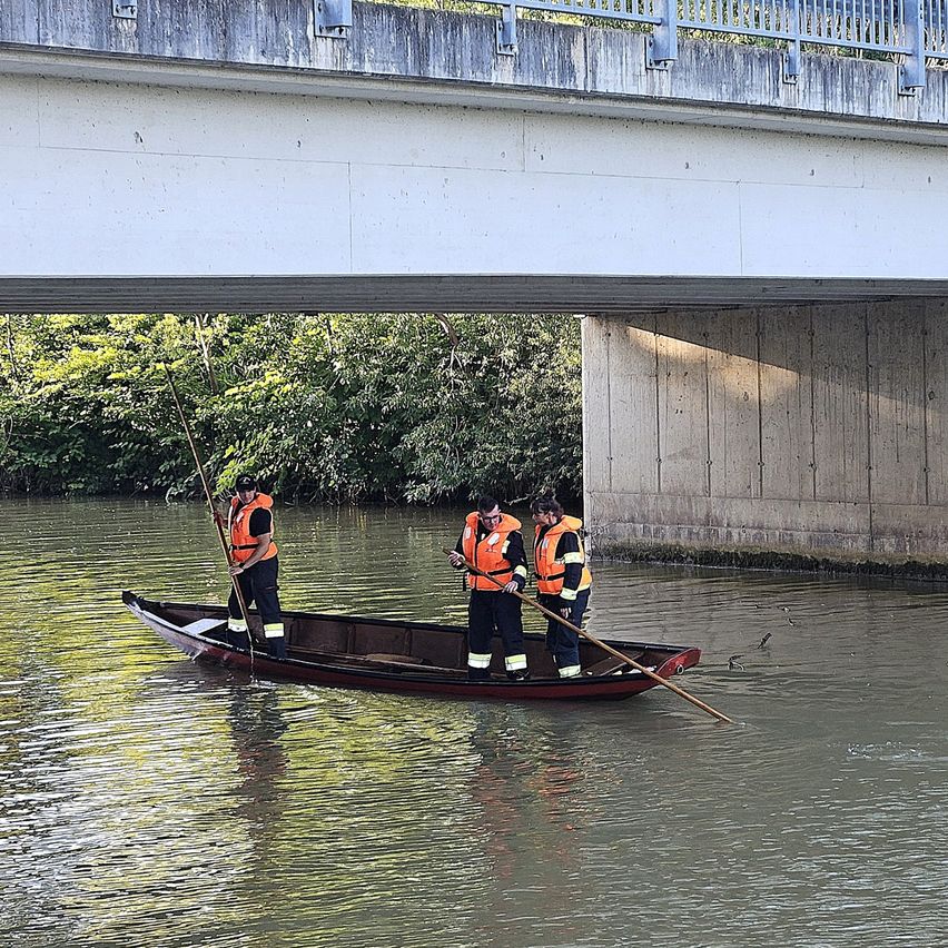 Drei Personen in Warnwesten paddeln ein Boot unter einer Brücke, einer benutzt einen langen Stab.