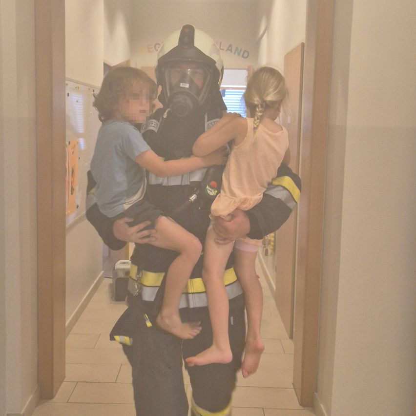 Ein Feuerwehrmann in Gasmaske und voller Uniform trägt zwei Kinder, einen Jungen und ein Mädchen, durch einen Flur.
