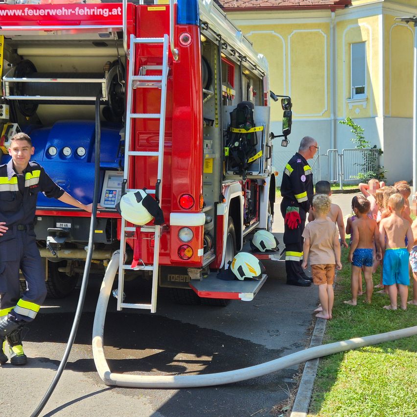 Zwei Feuerwehrleute erklären Kindern etwas über Brandschutz. Ein Feuerwehrmann hält eine Feuerwehrhose, der andere zeigt die Leiter des Feuerwehrwagens. Kinder stehen auf Gras, einige tragen Badekleidung.
