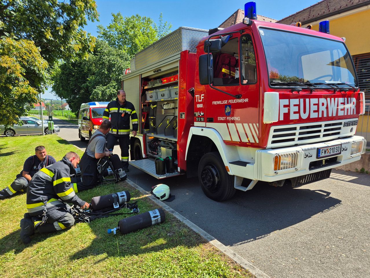 Ein Feuerwehrauto mit dem Wort Feuer ist am Straßenrand geparkt, wo drei Feuerwehrleute am Boden arbeiten.