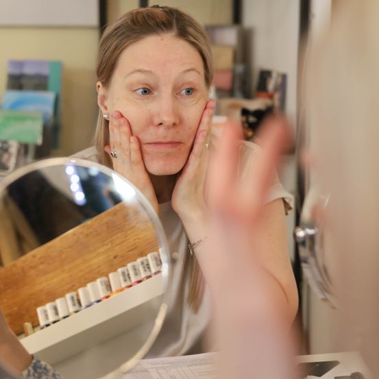 Eine Frau betrachtet ihr Gesicht im Spiegel. Sie hat ihre Hände auf den Wangen. Sie trägt ein weißes T-Shirt und ein Armband am Handgelenk. Im Spiegel ist die Reflexion eines Regals mit Nagellack zu sehen.