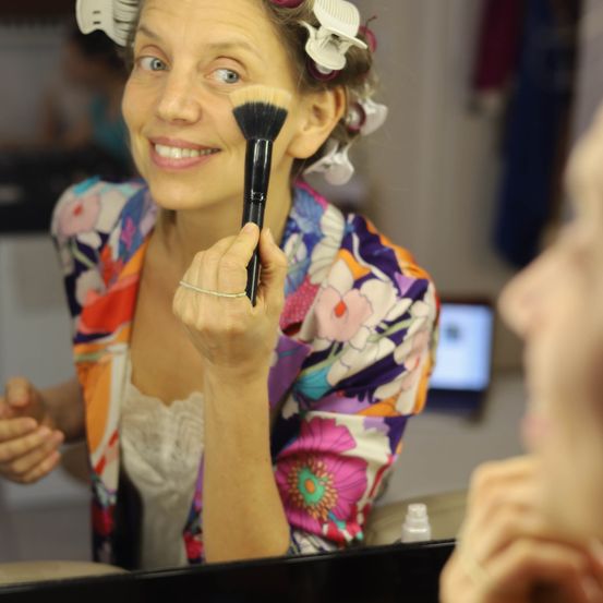 Eine Frau mit Lockenwicklern im Haar schminkt sich im Spiegel. Sie trägt eine farbenfrohe, florale Jacke und hält einen Make-up-Pinsel. Eine andere Person ist teilweise im Vordergrund zu sehen.