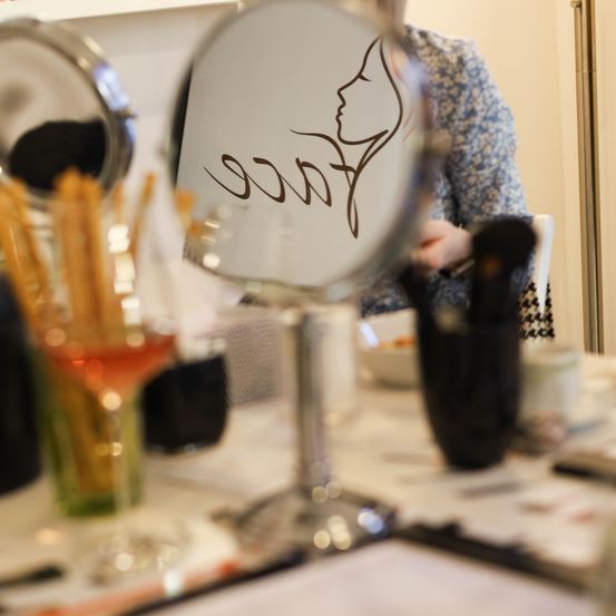 Ein Spiegel reflektiert das Gesicht einer Frau mit einer Zeichnung. In der Nähe sind Make-up-Pinsel und ein Weinglas auf einem Tisch.