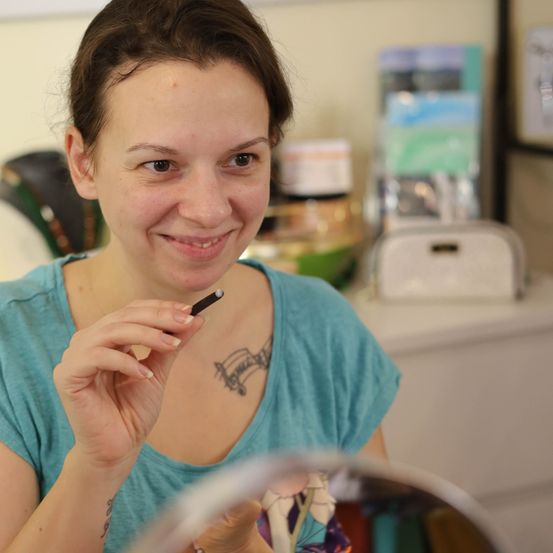 Eine Frau mit kurzen braunen Haaren hält einen Schminkstift, lächelt und schaut in einen Spiegel. Sie hat ein Tattoo auf der Brust und ist in einem Raum mit einem Schreibtisch und einem Toaster.