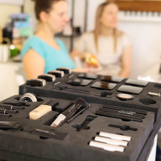 Zwei Frauen stehen hinter einem Tresen, eine hält Essen, die andere betrachtet verschiedene Make-up-Artikel in schwarzen Koffern.