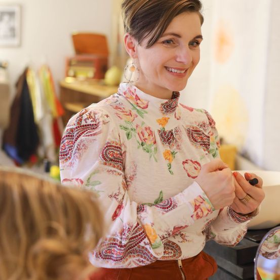 Eine lächelnde Frau in einem Blumenblusen schminkt ein Kind in einem Raum. Die Frau steht vor einem Spiegel und ein Kind sitzt vor ihr.