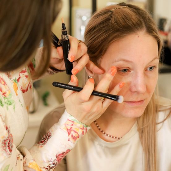 Eine Frau lässt sich von einer anderen Frau schminken, die einen Pinsel und einen Stift benutzt, um Make-up auf ihr Gesicht aufzutragen.