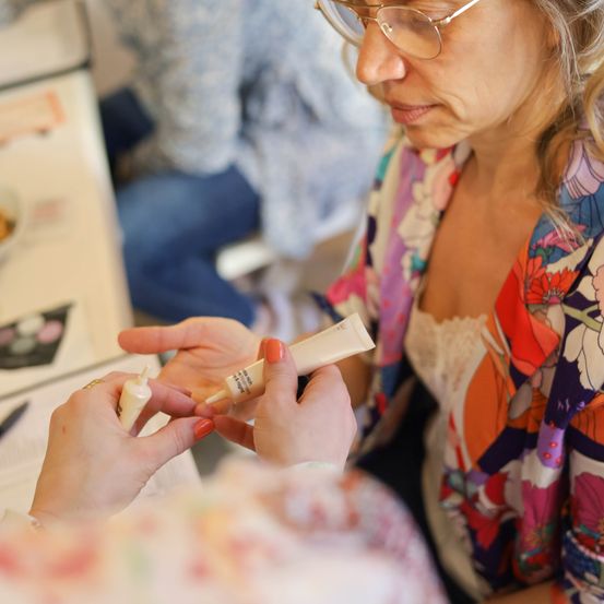 Eine ältere Frau mit Brille trägt jemandem Make-up auf, wobei sie sich auf deren Augen konzentriert.