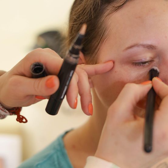 Eine Frau schminkt sich mit einem Stift und Pinsel. Sie hat orange Nägel und trägt ein blaues Hemd.