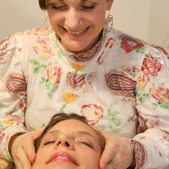 Eine Frau in einem Blumenhemd gibt einer anderen Frau, die mit geschlossenen Augen liegt, eine Gesichtsmassage.