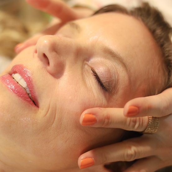 Eine Frau mit orangefarbenen Nägeln und rotem Lippenstift liegt auf dem Rücken und bekommt eine Gesichtsbehandlung. Ihre Augen sind geschlossen, und ihr Kopf wird von einer anderen Person gehalten.