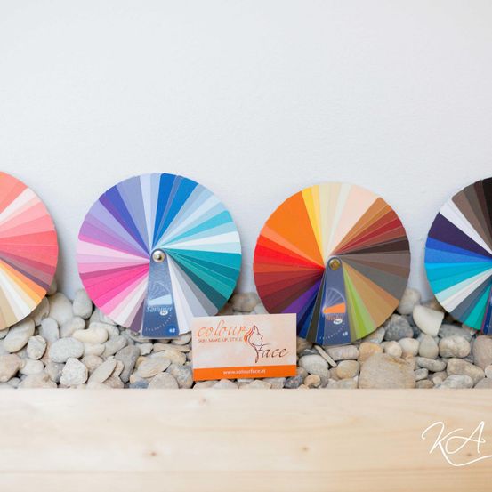 Eine Sammlung von Farbrädern mit verschiedenen Farbverläufen wird auf Kieselsteinen ausgestellt. Eine orangefarbene Visitenkarte mit Logo befindet sich in der Mitte.