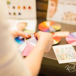Eine Person hält ein Farbrad in der Hand und betrachtet es genau. Verschiedene Farbmuster und eine Tabelle liegen auf dem Tisch vor ihnen. Im Hintergrund ist ein Plakat mit dem Gesicht einer Frau teilweise zu sehen.
