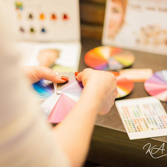 Eine Person hält ein Farbrad in der Hand und betrachtet es genau. Verschiedene Farbmuster und eine Tabelle liegen auf dem Tisch vor ihnen. Im Hintergrund ist ein Plakat mit dem Gesicht einer Frau teilweise zu sehen.