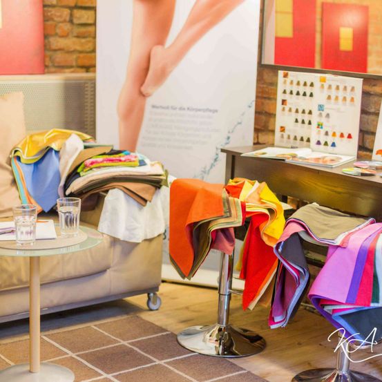 Ein Wohnzimmer mit einer Couch und einem Tisch, mehrere Hocker mit bunten Stoffen darüber gelegt und ein Holztisch mit verschiedenen Objekten. Ein Backsteinwand mit einem Poster im Hintergrund.