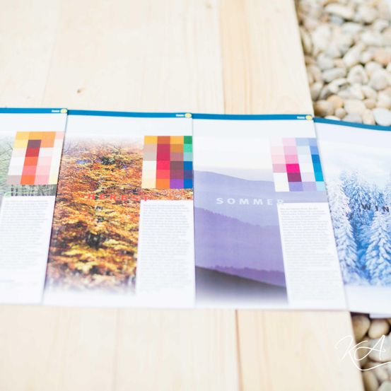 Eine Zeitschrift mit saisonalen Farbpaletten, einschließlich Herbst, Sommer und Winter, liegt auf einer hölzernen Oberfläche umgeben von Kieselsteinen.