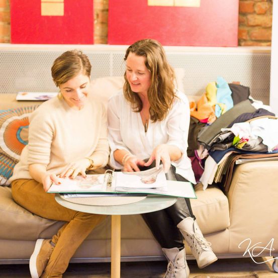 Zwei Frauen sitzen auf einer Couch, eine in einem weißen Shirt und die andere in einem cremefarbenen Pullover, und betrachten ein Fotoalbum auf einem Tisch vor ihnen.