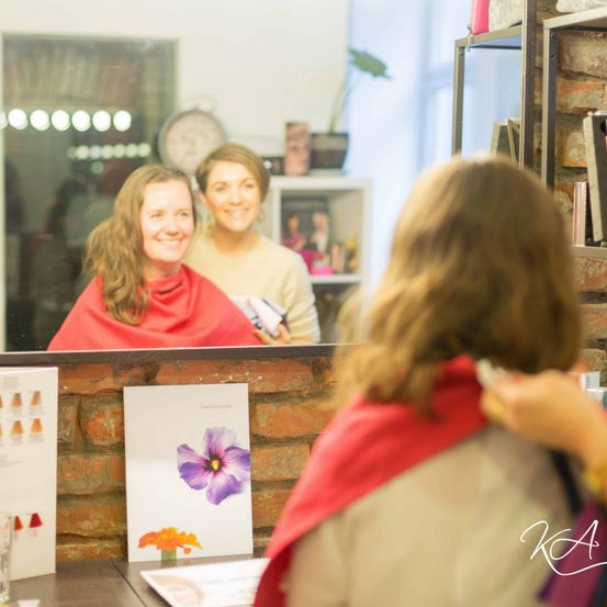 Eine Frau lässt sich in einem Salon die Haare stylen, während eine andere Frau im Spiegel lächelt. Die Wand dahinter hat ein Ziegelmuster und ein Poster.