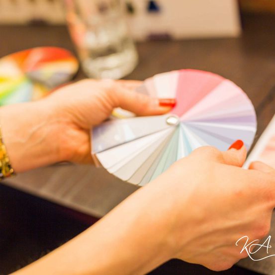 Eine Person hält ein Farbrad mit den Fingern am Rand und roten Nägeln. Ein verschwommenes Glas steht auf einem Tisch. Ein farbenfroher Teller ist links.