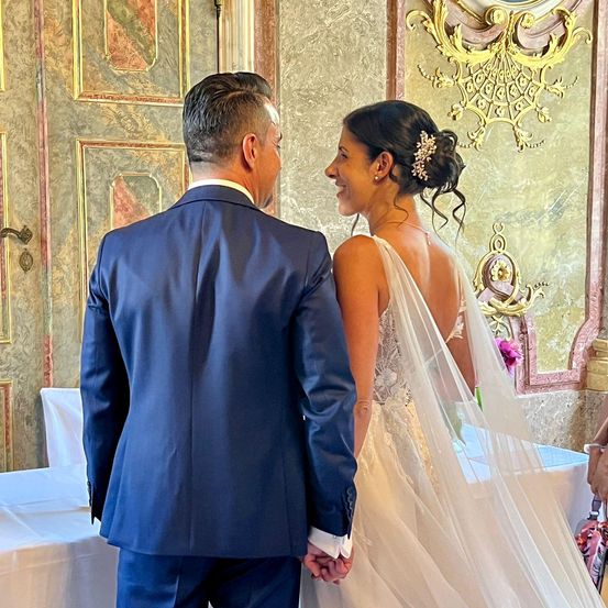 Ein Mann und eine Frau stehen in einem Raum, möglicherweise ein Hochzeitssaal. Der Mann trägt einen blauen Anzug, während die Frau ein Hochzeitskleid und Schleier trägt. Sie scheinen einen zärtlichen Moment zu teilen.