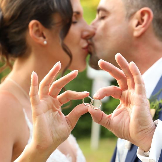 Eine Braut und ein Bräutigam halten zwei Eheringe zwischen den Fingern und küssen sich im Freien.