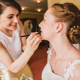 Eine Frau schminkt eine andere Frau, die ein Hochzeitskleid und eine Perlenkette trägt.