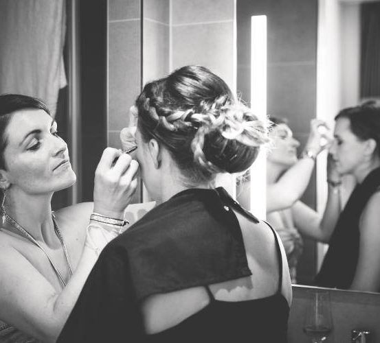 Eine Frau schminkt eine andere Frau im Spiegel, wobei zwei weitere Frauen im Hintergrund zu sehen sind. Der Raum ist schwach beleuchtet und die Wände sind gefliest.