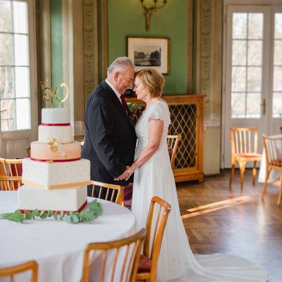 Ein älteres Paar steht neben einem Tisch mit einem großen dreistöckigen Kuchen mit goldenen Zahlen 50 obenauf. Der Raum hat grüne Wände und Holzmöbel.