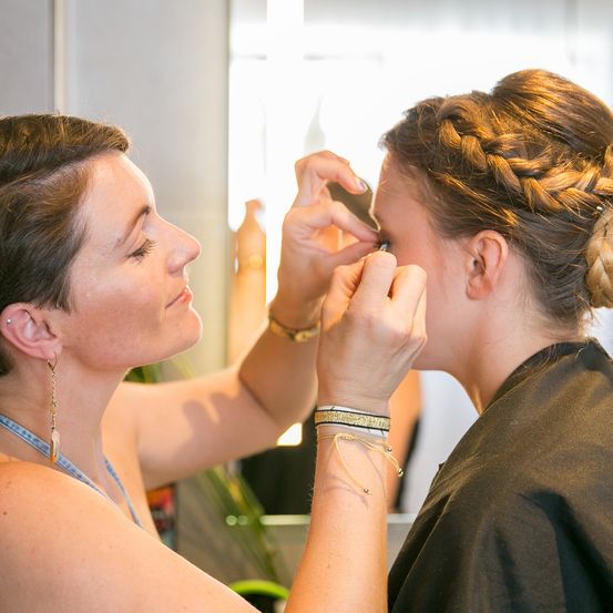 Eine Frau schminkt das Gesicht einer anderen Frau und konzentriert sich auf ihre Augen. Die Frau, die Make-up aufträgt, hat eine geflochtene Frisur. Sie befinden sich in einem Raum mit einem Spiegel.