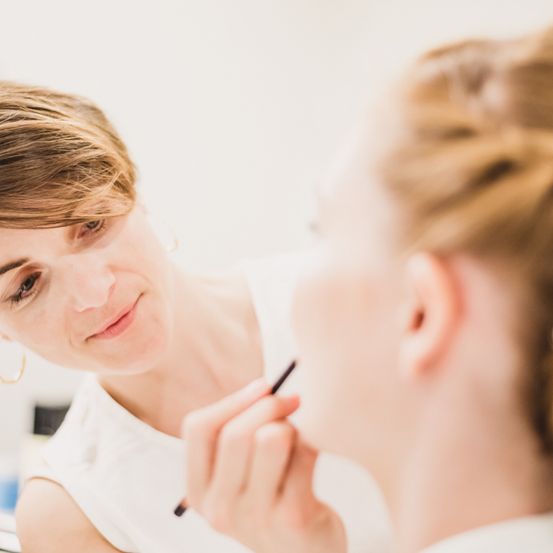 Eine Frau schminkt das Gesicht einer anderen Frau in einem weißen Raum, die Visagistin lächelt.