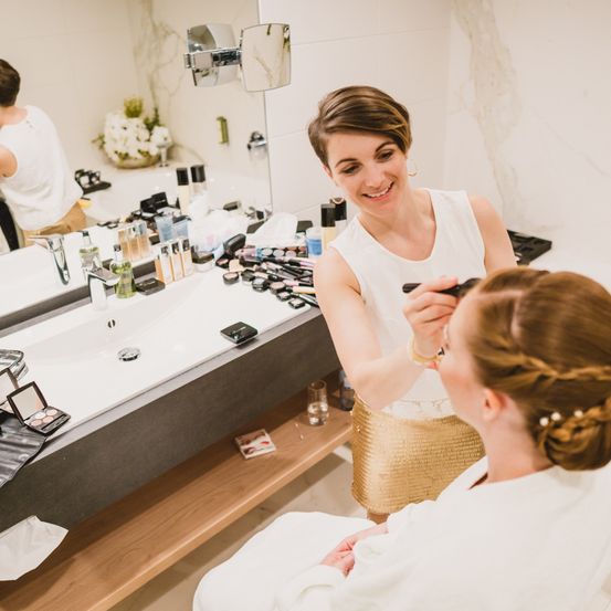 Eine Frau lässt sich von einer anderen Frau im Badezimmer das Make-up schminken. Auf dem Tresen liegen Make-up-Produkte.