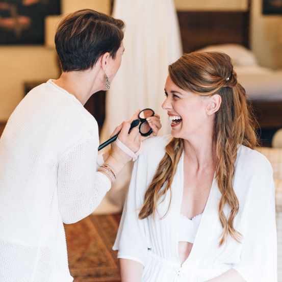 Eine Frau lacht und lässt sich von einer anderen Frau, die einen Pinsel hält, das Make-up machen.