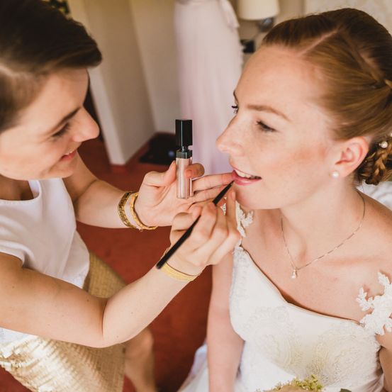 Eine Visagistin schminkt eine Braut, die nach unten schaut, in einem weißen Kleid und einer Halskette.