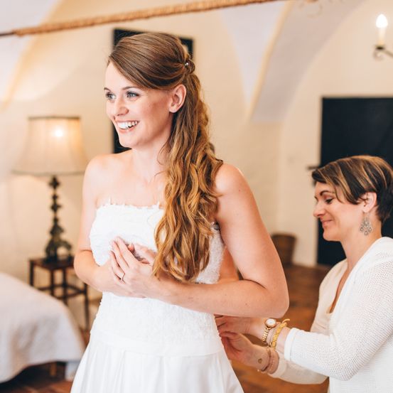 Eine Braut in einem weißen Kleid lächelt, während eine Frau ihr Kleid in einem Raum mit warmem Licht und rustikalem Dekor anpasst.