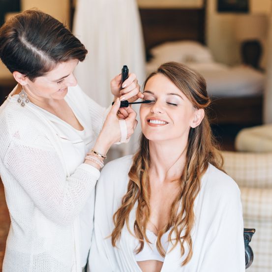 Eine Frau trägt Eyeliner auf die Augen einer anderen Frau auf, die mit geschlossenen Augen lächelt. Hinter ihnen befindet sich ein Bett mit Kissen und eine Wand mit einem Bilderrahmen.