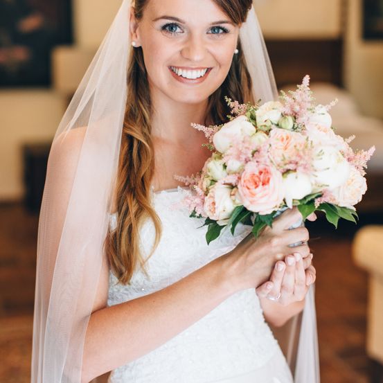 Eine Braut in einem weißen Kleid und Schleier hält einen Strauß aus rosa und weißen Blumen und lächelt in einem Raum.