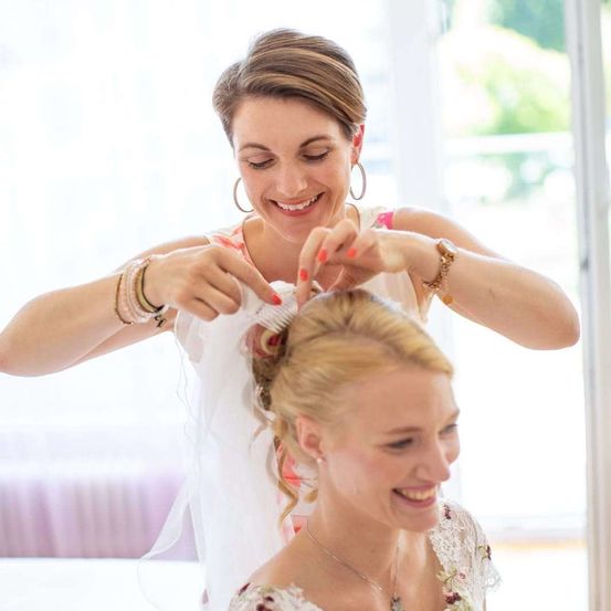 Eine Frau mit kurzen Haaren fixiert das Haar einer Braut in einem weißen Kleid. Beide lächeln und sehen sich an.